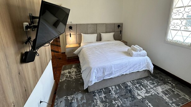 A hotel room with a bed, a TV mounted on the wall, a nightstand, a lamp, and a window with decorative iron bars.