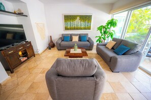 A living room with a grey sofa set, a coffee table, a TV, and a large window with a view of greenery.