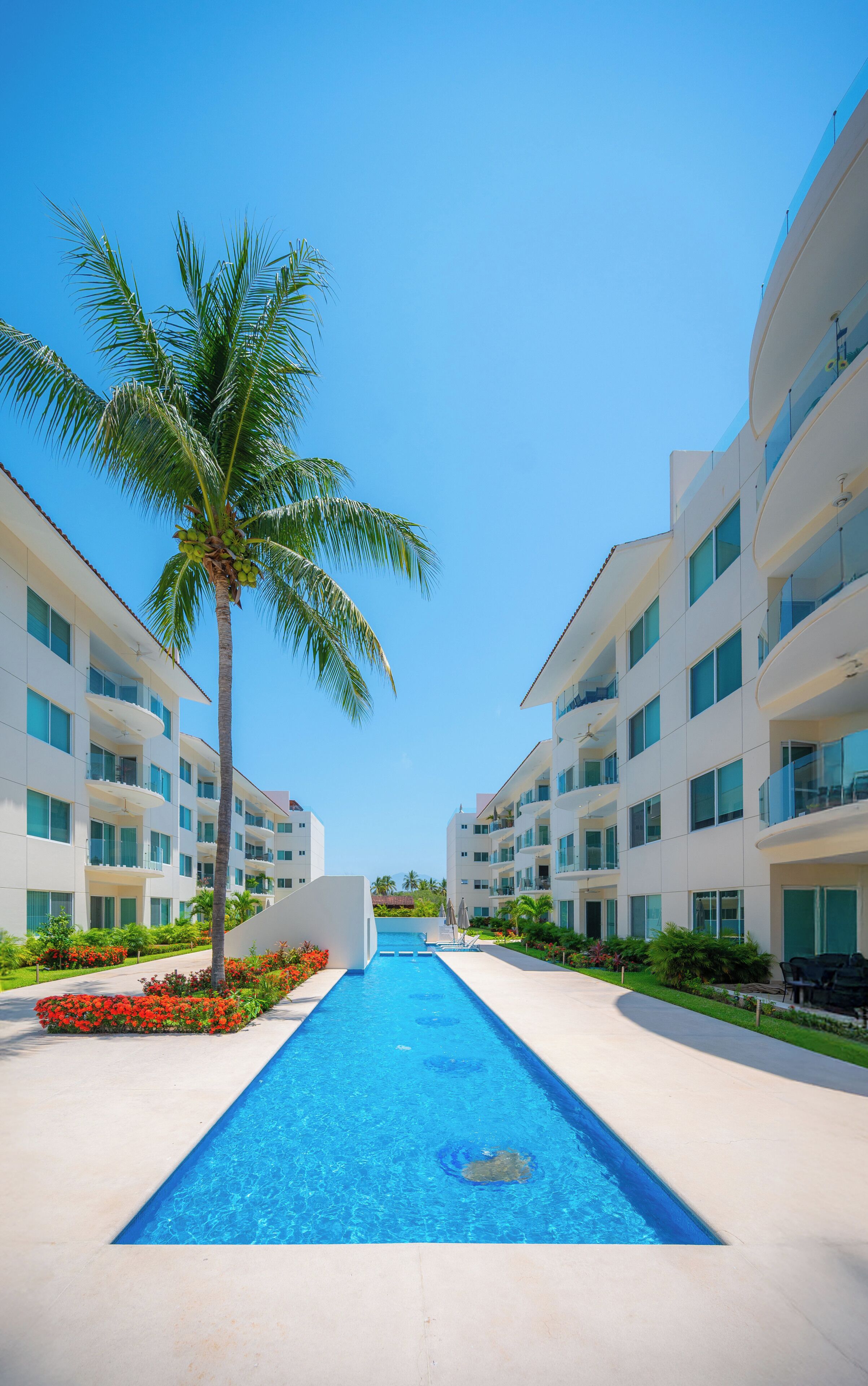 A modern hotel with a central swimming pool, surrounded by palm trees and a clear blue sky.