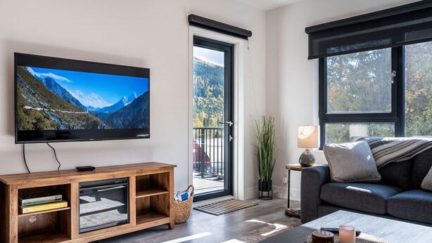 A modern living room with a television, a sofa, a coffee table, and a view of a mountainous landscape through a sliding door. - Mountainview Escape Revelstoke Vacations (Revelstoke)