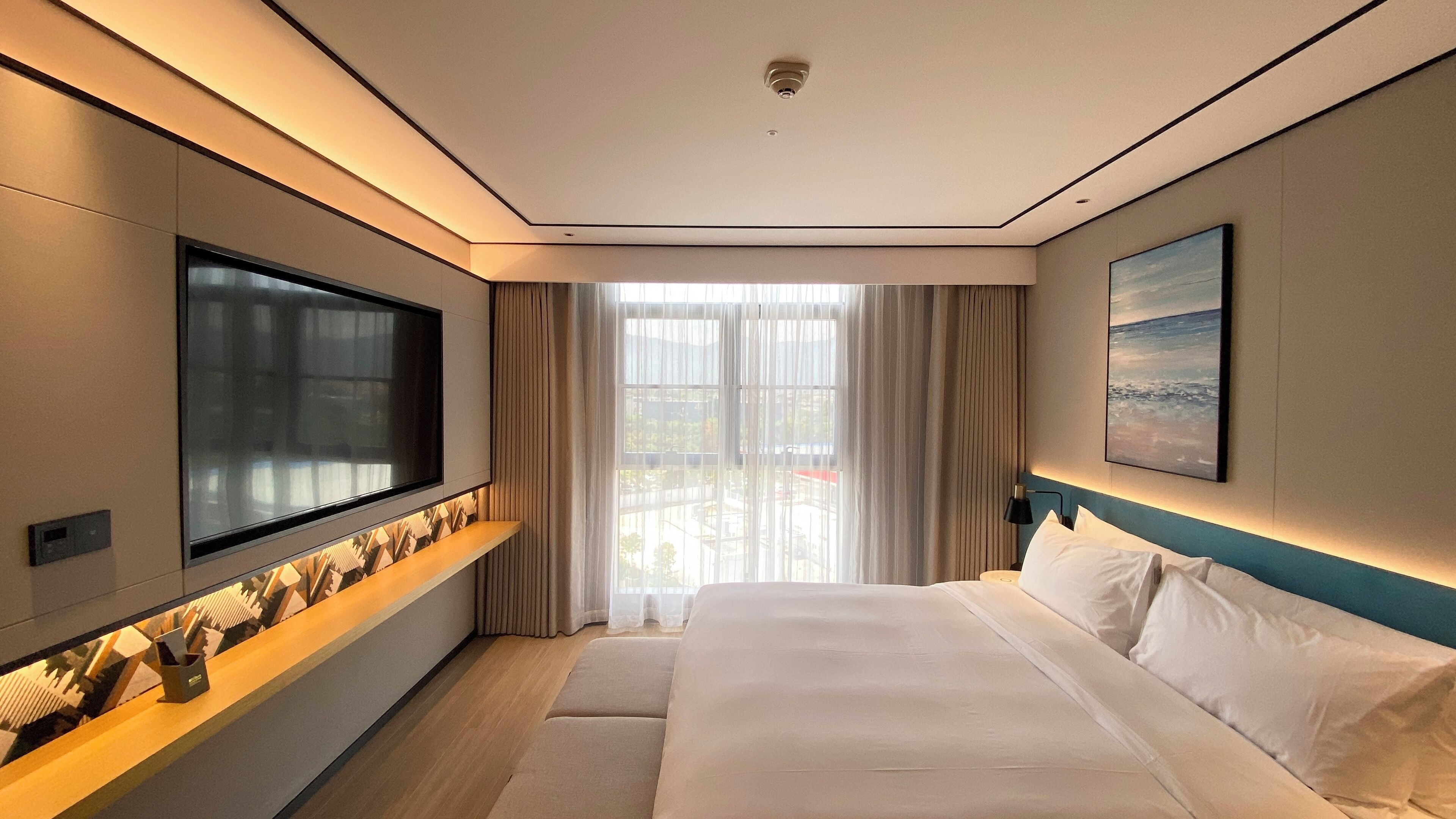 Una habitación de hotel moderna con un televisor de pantalla plana grande montado en la pared, una cama con ropa blanca, vistas a la ciudad a través de ventanas amplias y un cuadro en la pared.