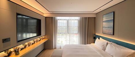 Una habitación de hotel moderna con un televisor de pantalla plana grande montado en la pared, una cama con ropa blanca, vistas a la ciudad a través de ventanas amplias y un cuadro en la pared.