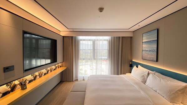 Una habitación de hotel moderna con un televisor de pantalla plana grande montado en la pared, una cama con ropa blanca, vistas a la ciudad a través de ventanas amplias y un cuadro en la pared.