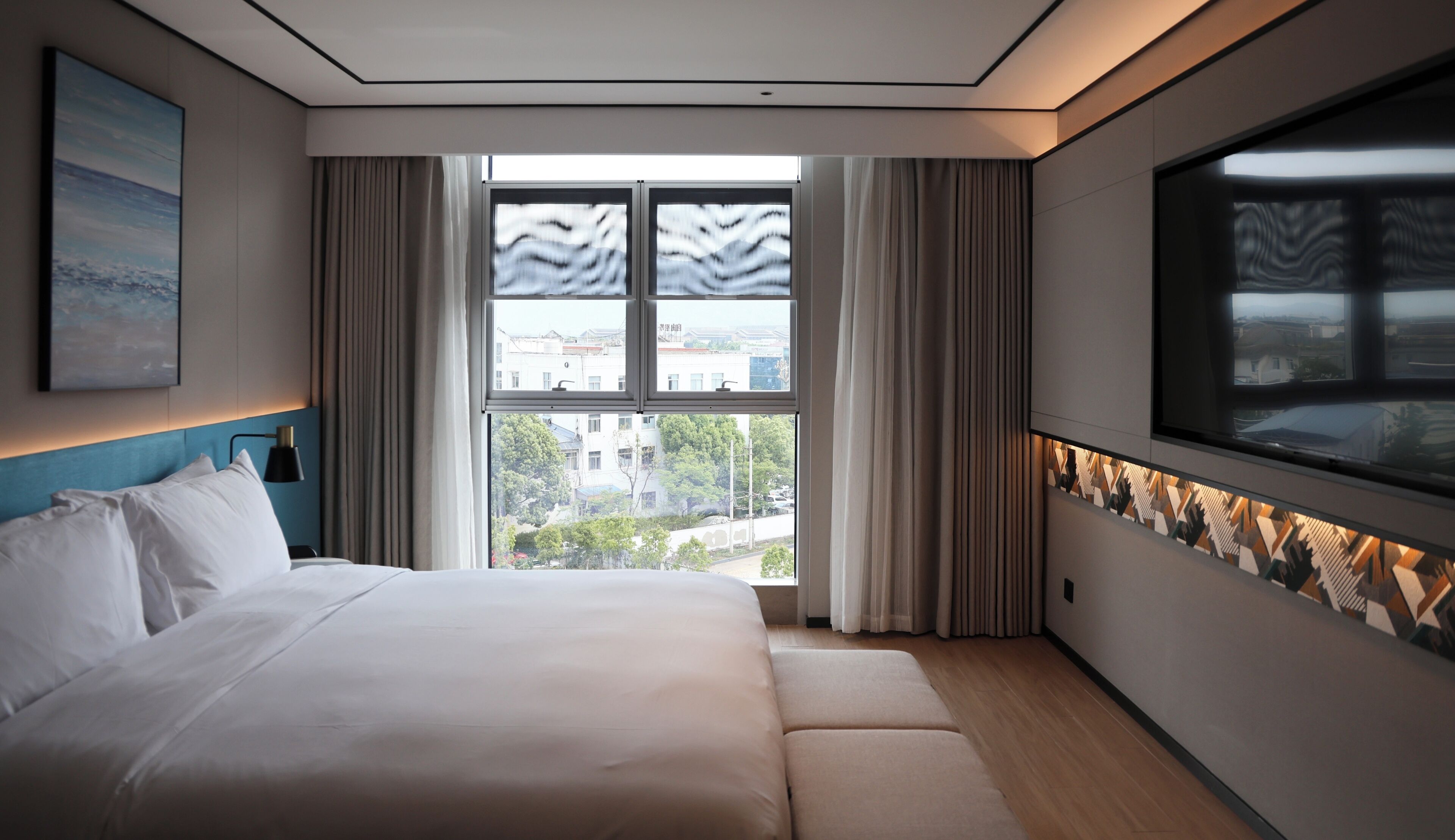 A modern hotel room with a large bed, a view of the city through floor-to-ceiling windows, and a decorative wall panel.