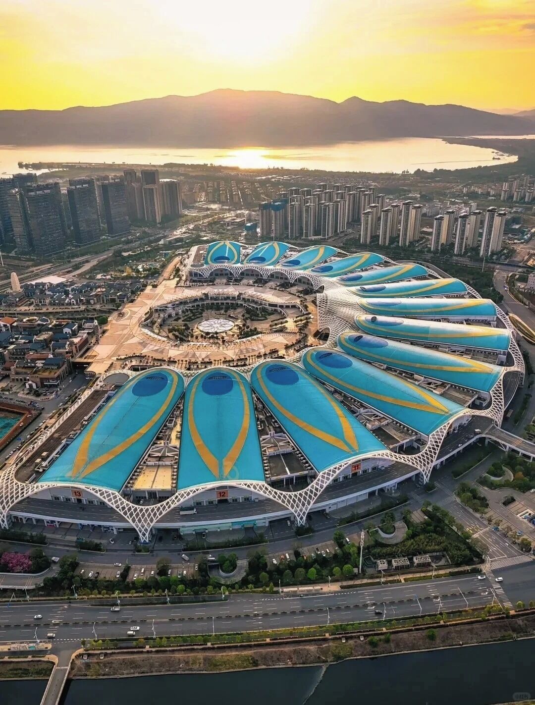 A large stadium with multiple blue and yellow roof sections, surrounded by high-rise buildings and a body of water.