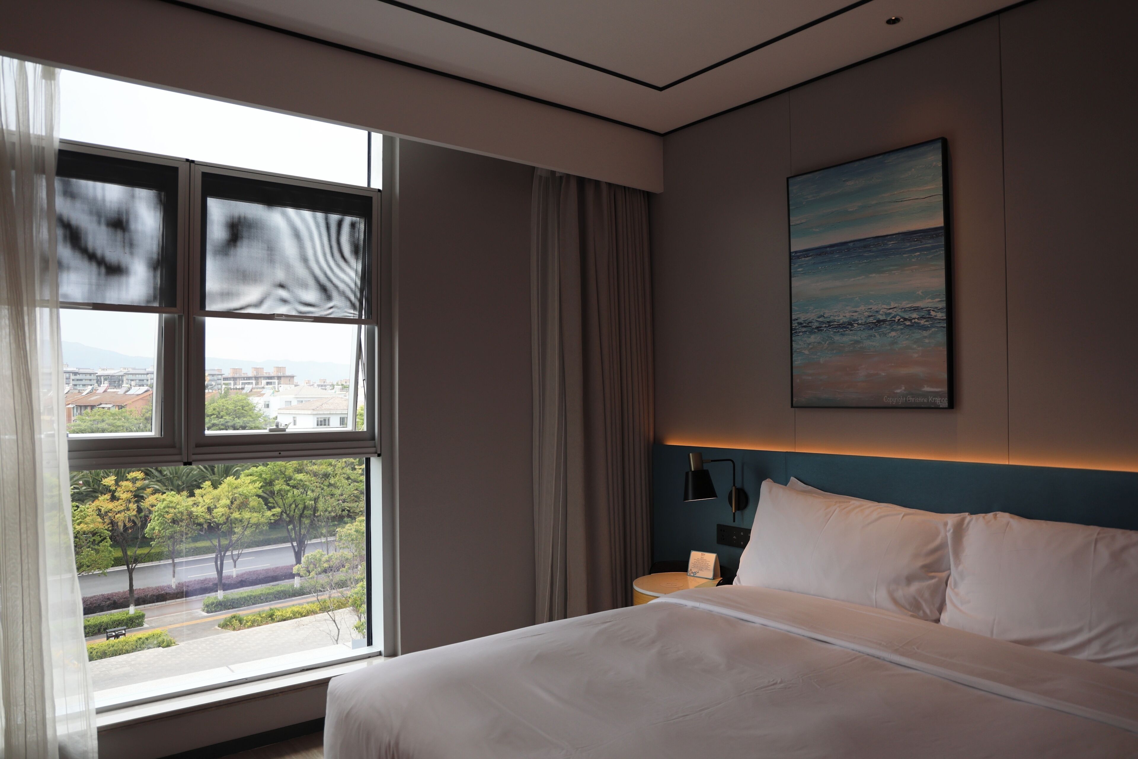 A hotel room with a large window, a bed, a nightstand, and a painting on the wall.