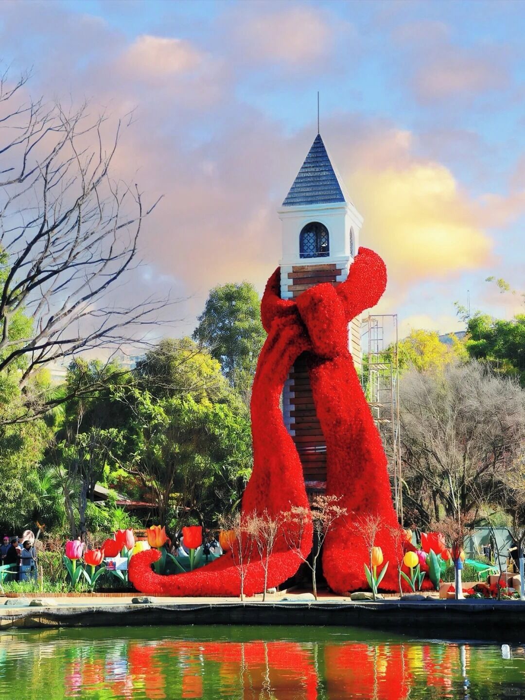 Eine große, rote Skulptur, die eine Figur mit einem Turm darstellt, umgeben von einem Garten mit verschiedenen Pflanzen und einem Teich.