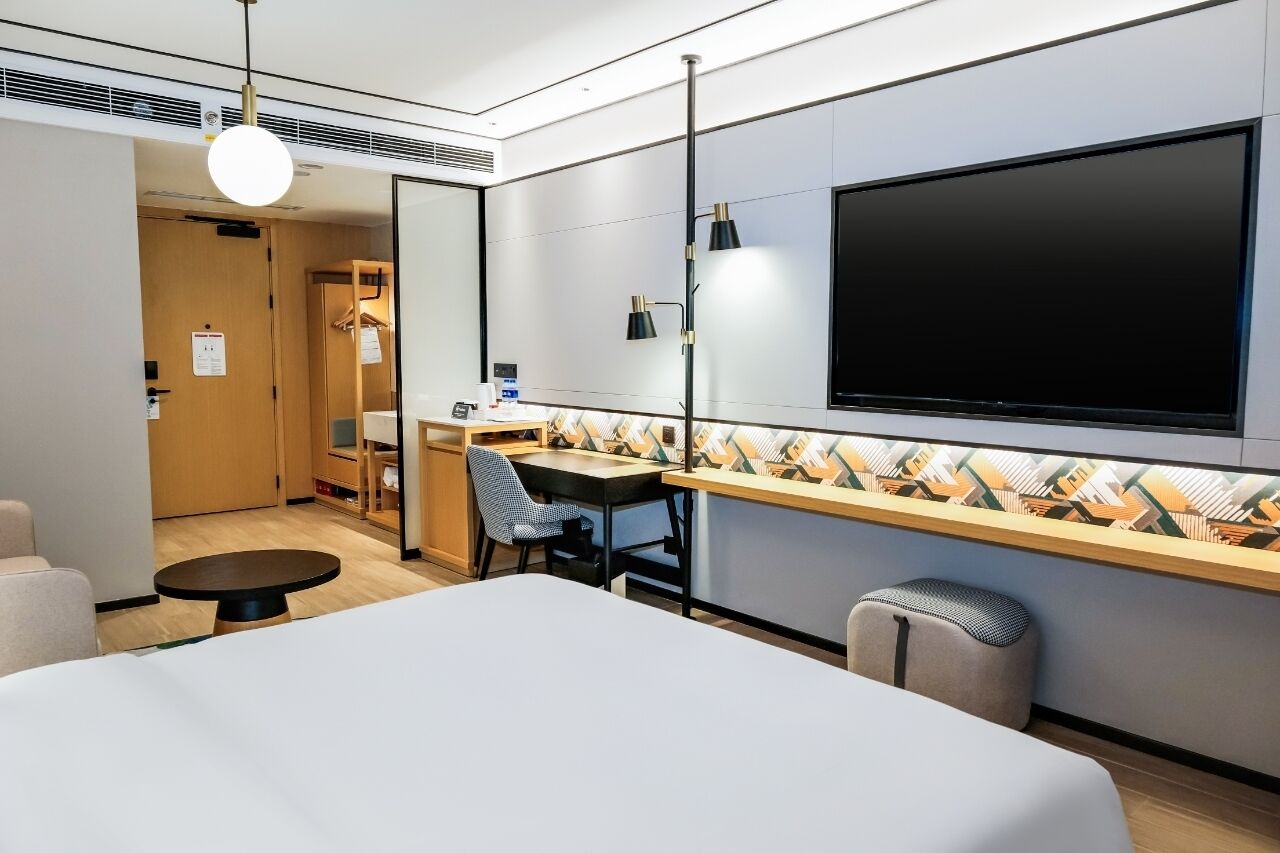 A modern hotel room with a bed, a desk, a chair, a TV, and a small stool.