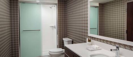 A modern bathroom with a glass shower enclosure, a white toilet, a sink with a mirror, and patterned wallpaper.