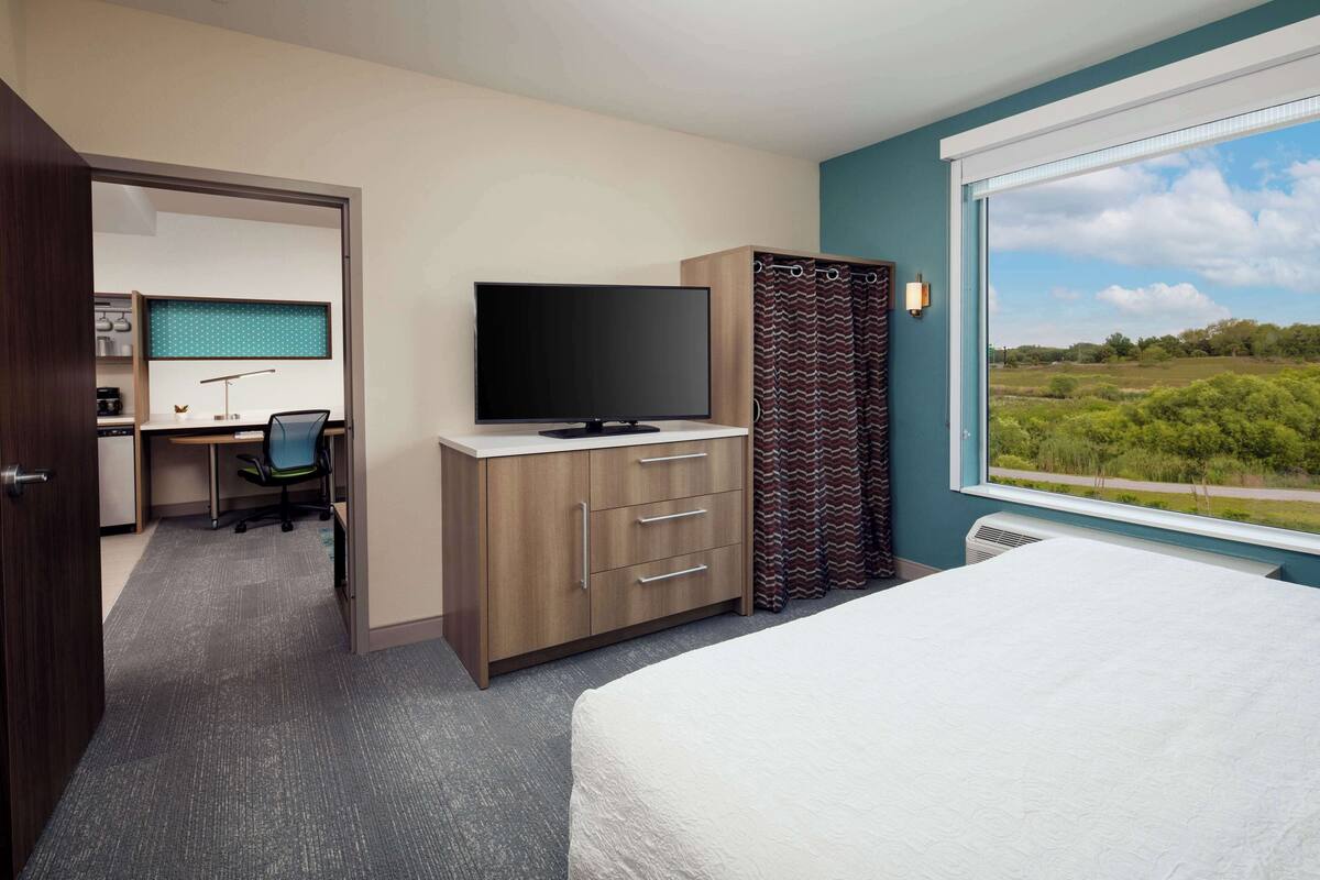 A hotel room with a bed, a desk with a computer, a TV, and a window with a view of greenery.