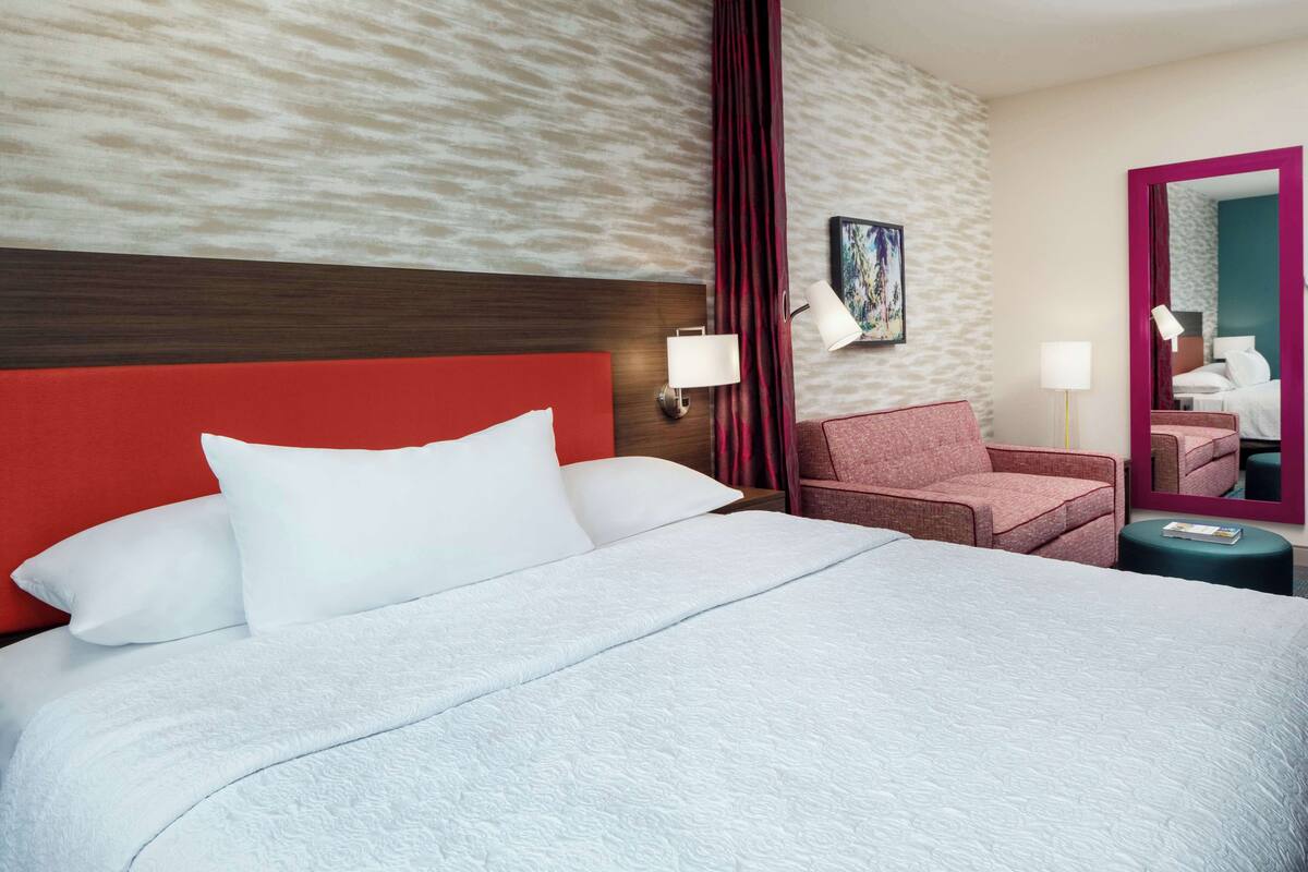A hotel room with a large bed, a red headboard, a pink armchair, a small table, and a mirror.