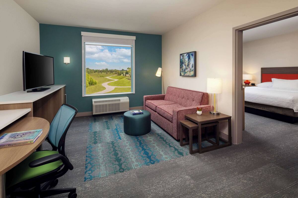 A hotel room with a desk, chair, TV, sofa, ottoman, lamp, and a bed.