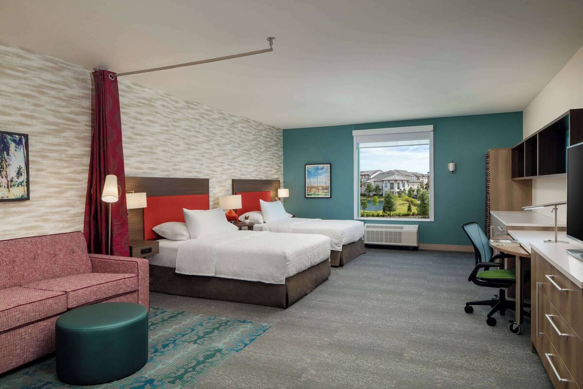 A hotel room with two beds, a desk, a chair, a sofa, and a view of a building through a window.
