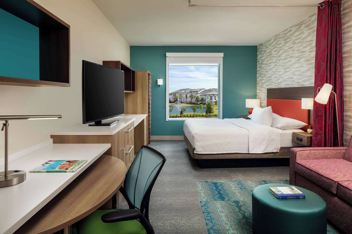 A hotel room with a bed, desk, chair, television, and a view of buildings through a window.