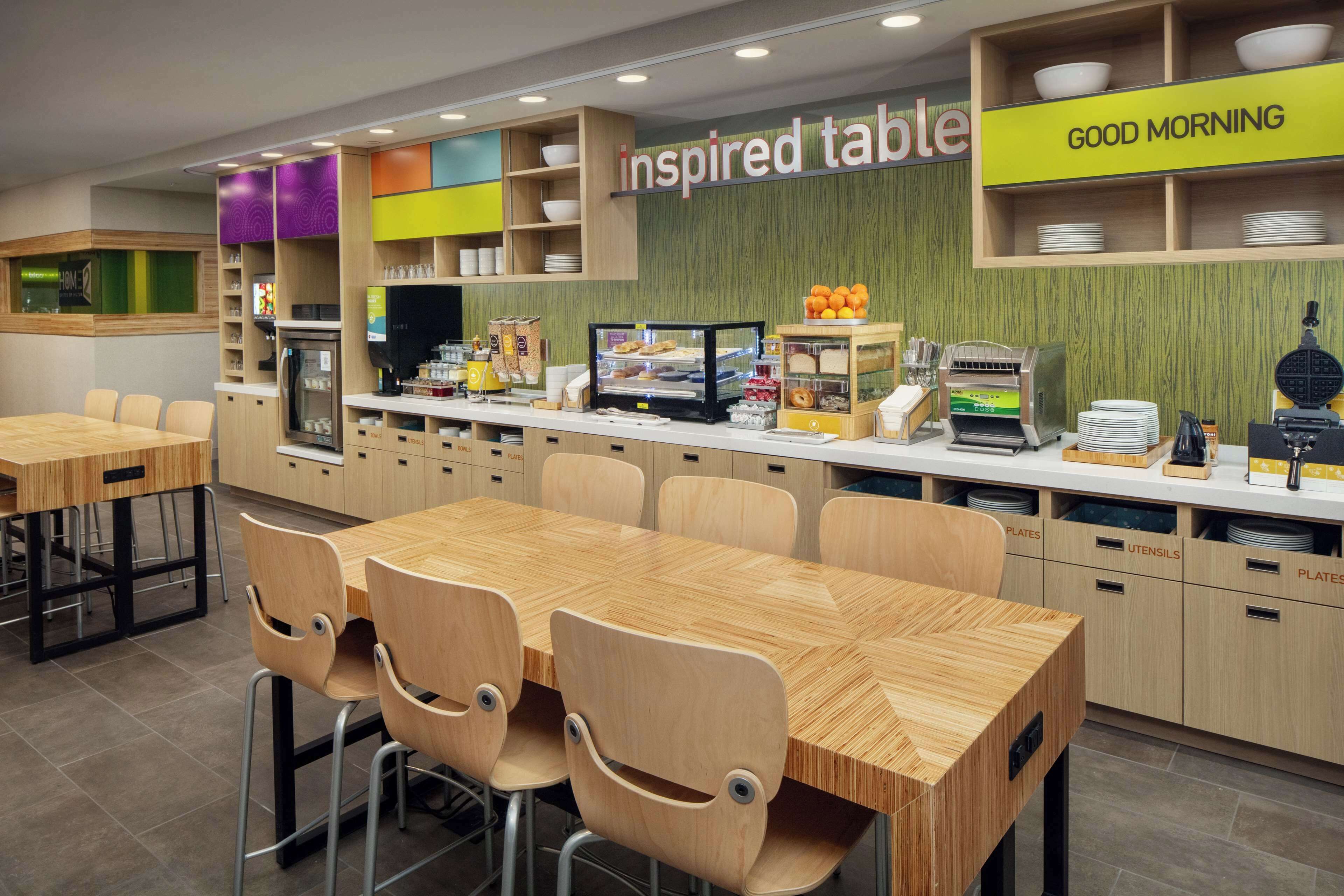 Un'area colazione moderna con tavoli e sedie in legno, una vetrina con prodotti alimentari e un cartello con la scritta "inspired table".