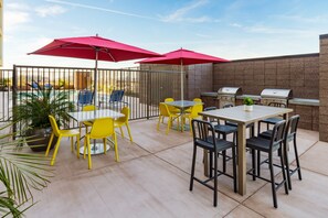Un patio al aire libre con sillas amarillas y negras, sombrillas rojas y una parrilla de barbacoa.