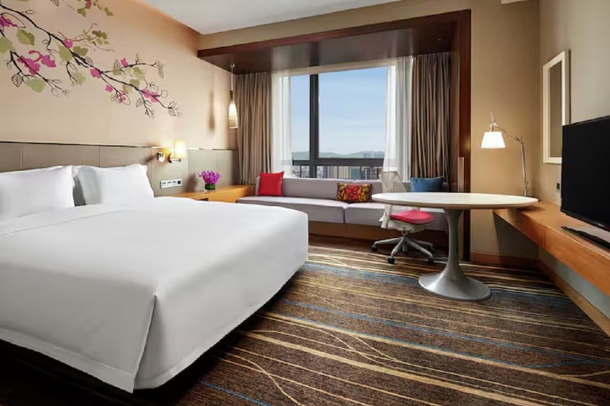 Habitación de hotel con una cama grande, una mesita redonda pequeña, un sofá con cojines, un televisor y una ventana con cortinas.