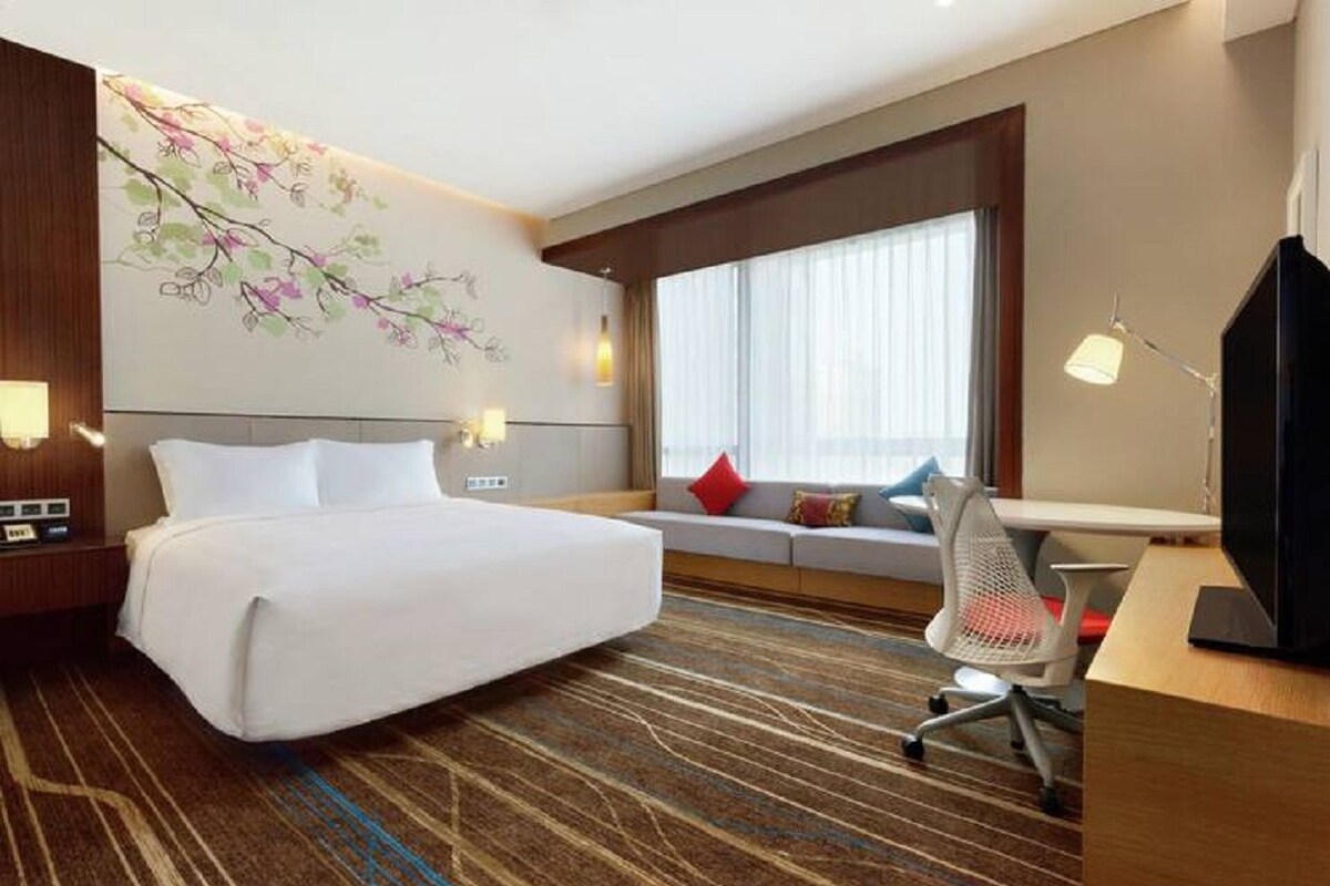 Habitación de hotel con una cama grande, un escritorio con una silla, un televisor, un sofá con cojines y una pared con un mural floral.