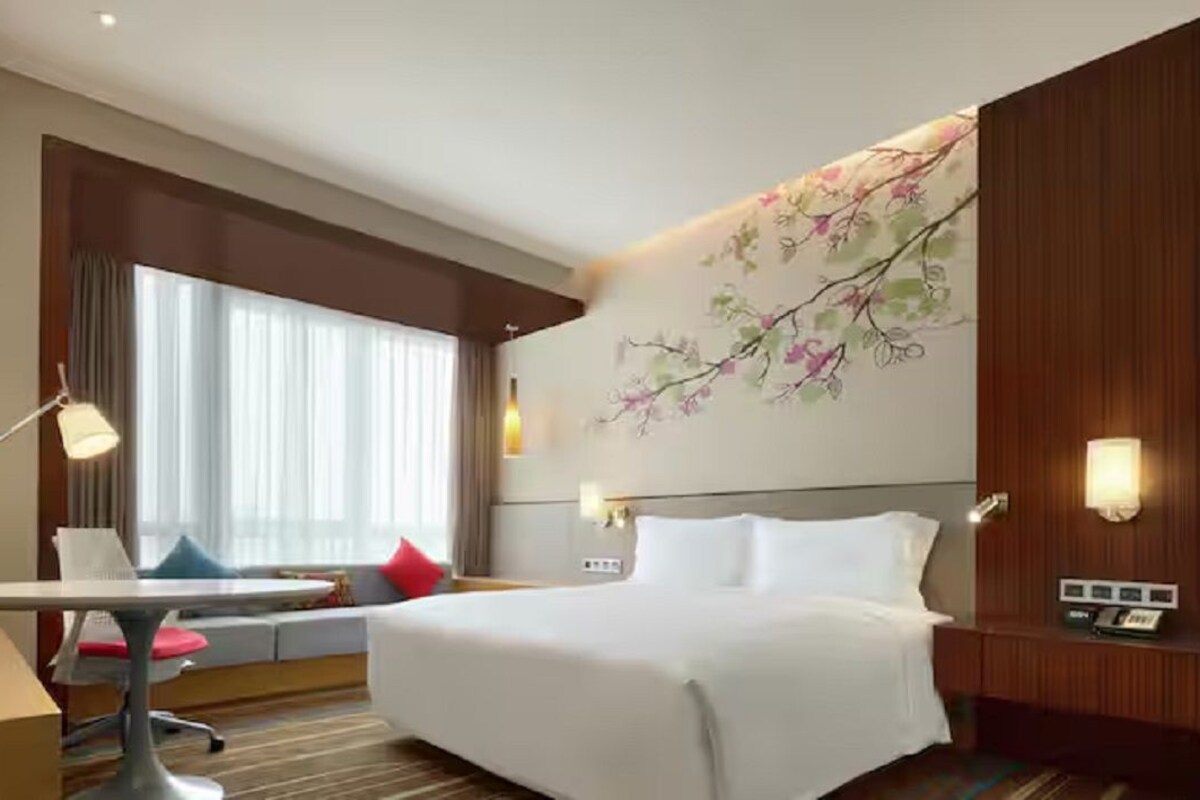 Una habitación de hotel con una cama, un escritorio con silla, un televisor, una ventana con cortinas y un mural floral en la pared.