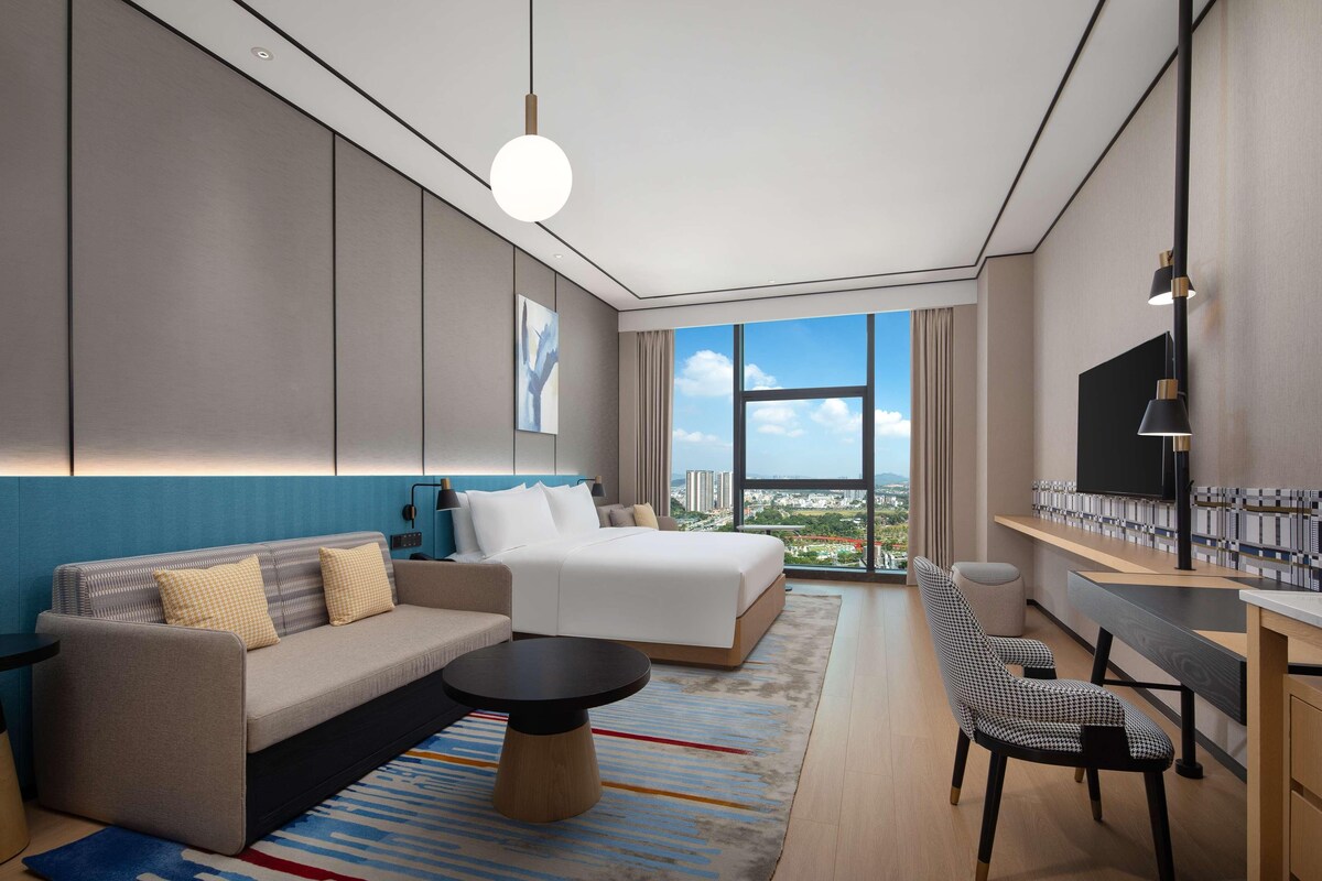 Habitación de hotel moderna con una cama grande, un sofá, una mesa de comedor y vistas a la ciudad.