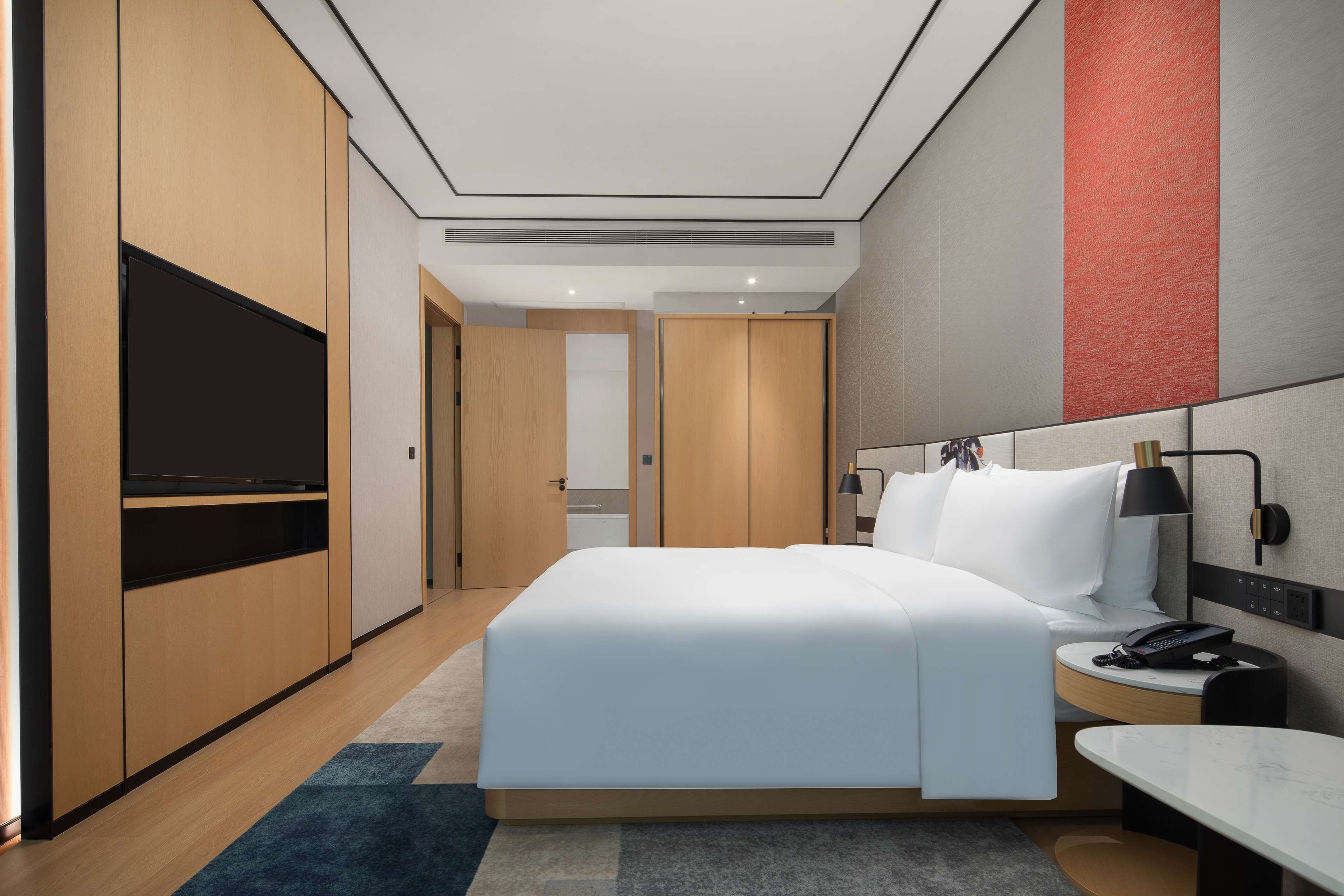 Une chambre d’hôtel moderne avec un grand lit, un téléviseur posé sur un meuble, une table de chevet avec une lampe et un bureau avec un téléphone.