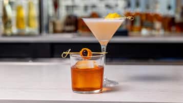 Two cocktails on a bar counter with a blurred background of bottles.
