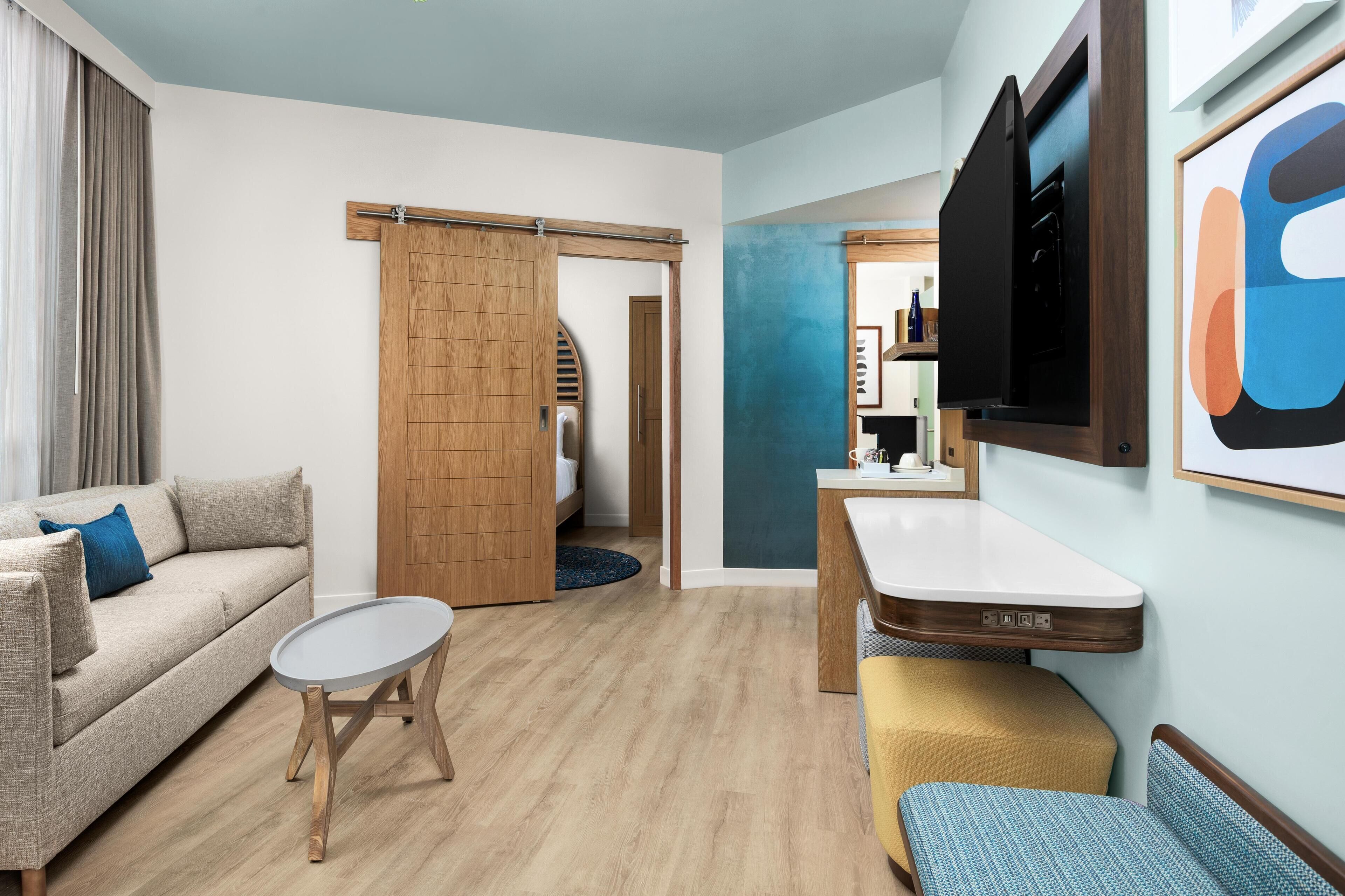 Habitación de hotel compacta con sofá, una pequeña mesa redonda, una banca, televisor y una puerta corredera que da a un baño.