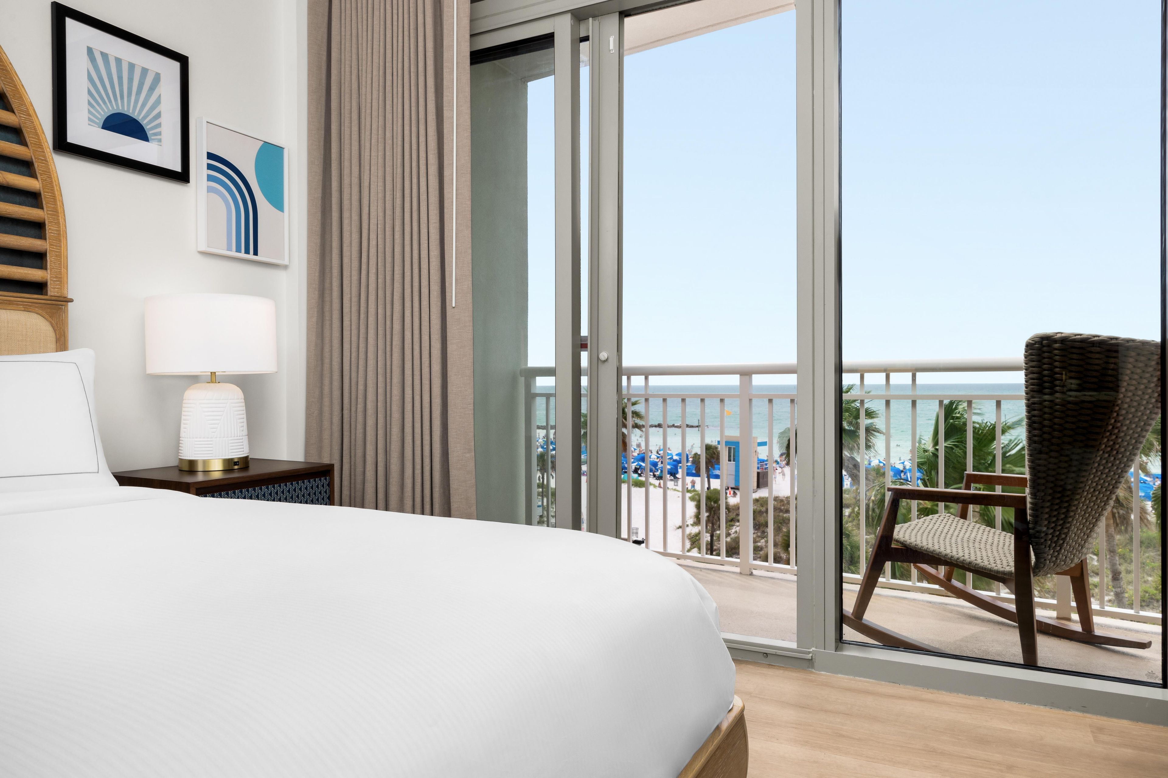 Ein Hotelzimmer mit Bett, Nachttisch, Lampe, Stuhl und einem Balkon mit Blick auf den Strand.