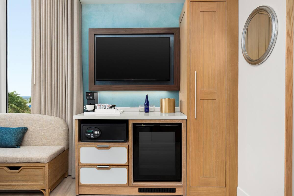 Een moderne kamer met een flatscreen televisie bevestigd aan een houten kast, een compacte koelkast, een koffiezetapparaat, en een klein tafeltje met een kop en een fles.
