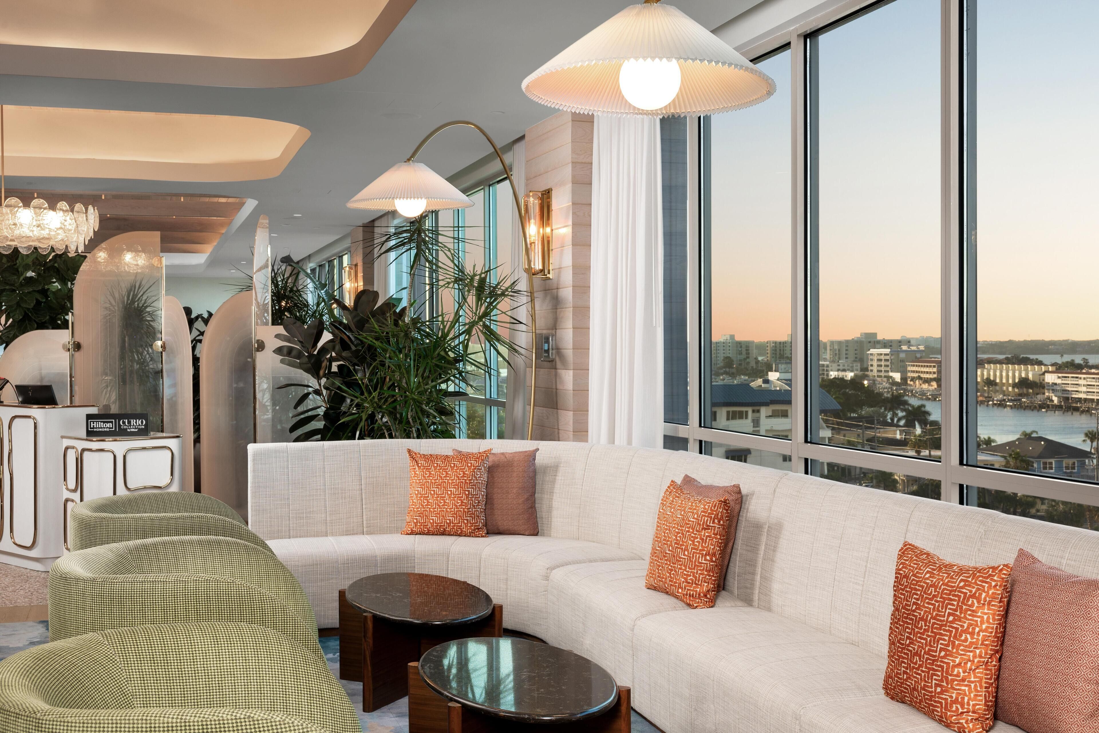 Un hall d’hôtel moderne avec un canapé blanc, des fauteuils verts et une grande fenêtre donnant sur la ville.