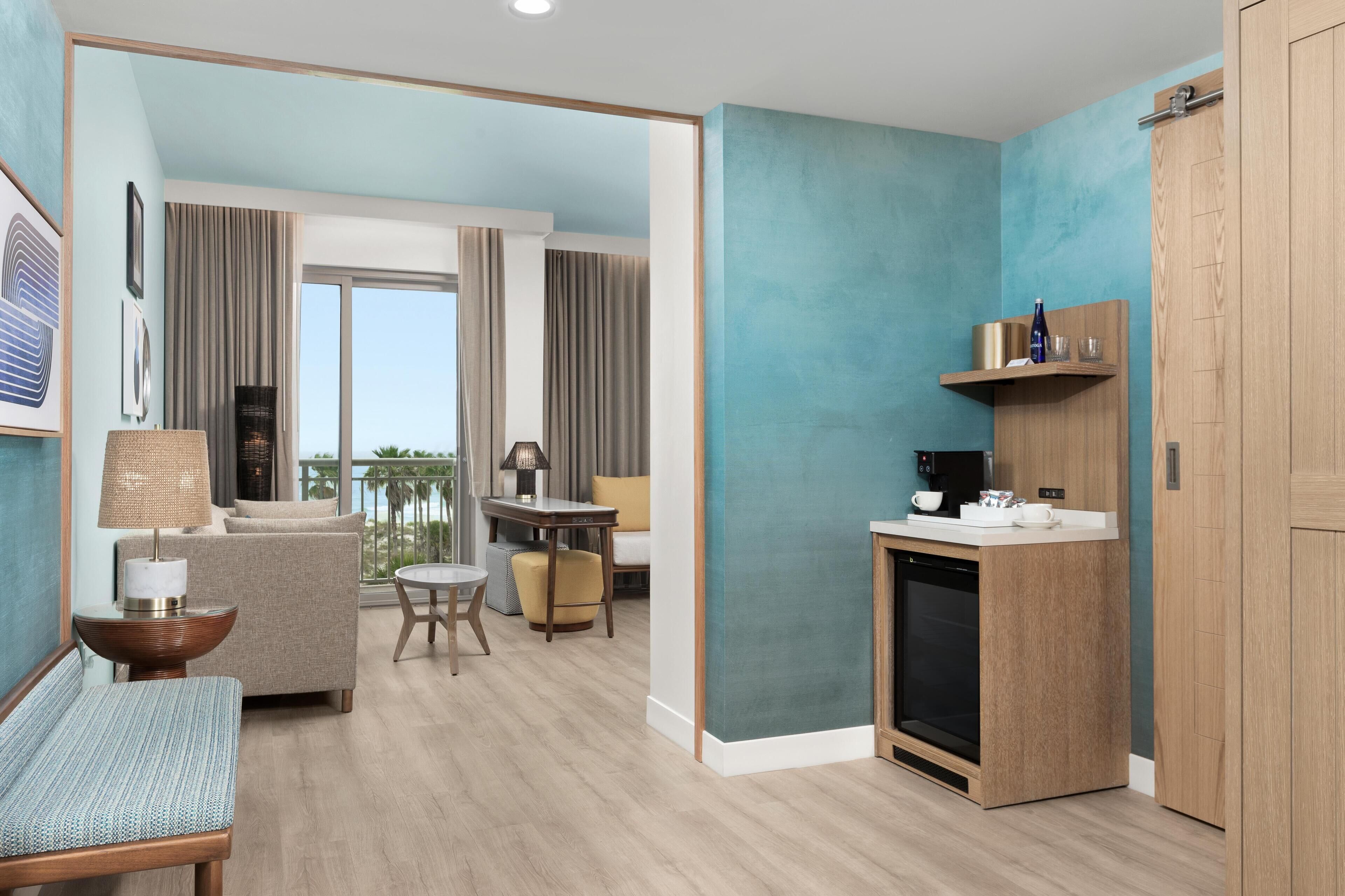 Una habitación de hotel moderna con balcón, kitchenette y zona de estar con sofá y una pequeña mesa.