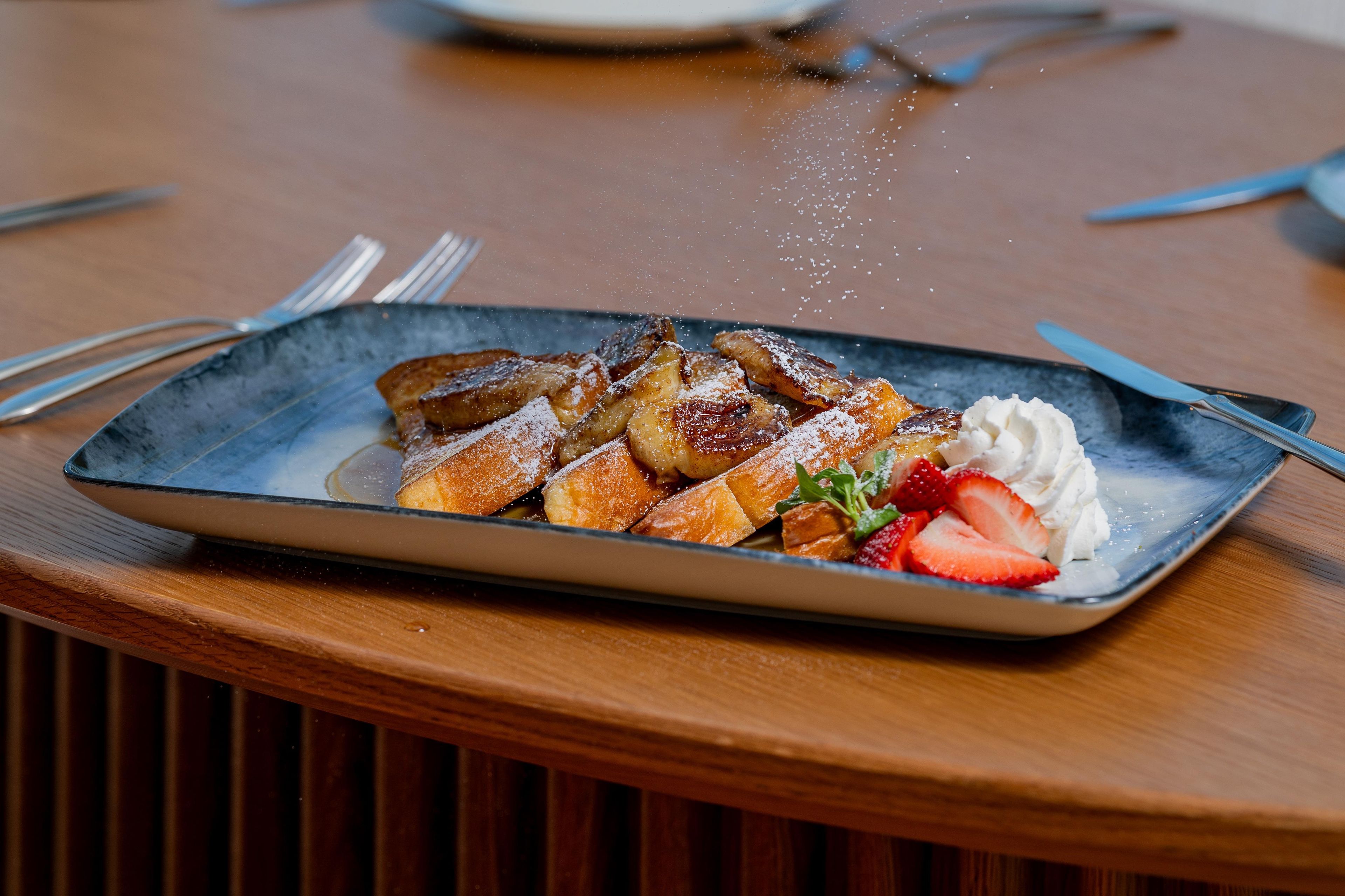 Un plato de comida con un postre, posiblemente pudín de pan, decorado con fresas y crema batida, sobre una mesa de madera.