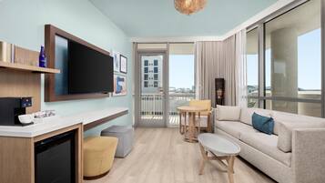 A modern living room with a sofa, coffee table, and TV. There is a balcony with a view of buildings.