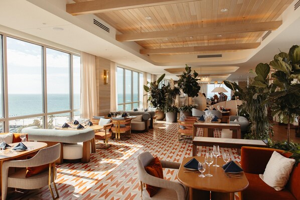 A spacious lounge area with large windows overlooking the sea, furnished with sofas, tables, and chairs, and decorated with potted plants.