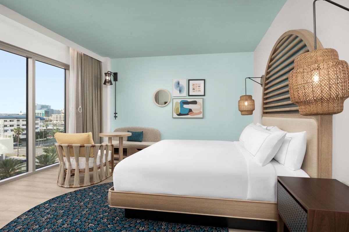 Een moderne hotelkamer met een groot bed, een zithoek met een stoel en tafel, een groot raam met uitzicht op de stad, en decoratieve muurkunst.