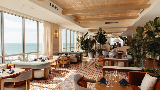 Een ruime, goed verlichte lounge met grote ramen met uitzicht op zee, comfortabele zitplaatsen en veel groen.