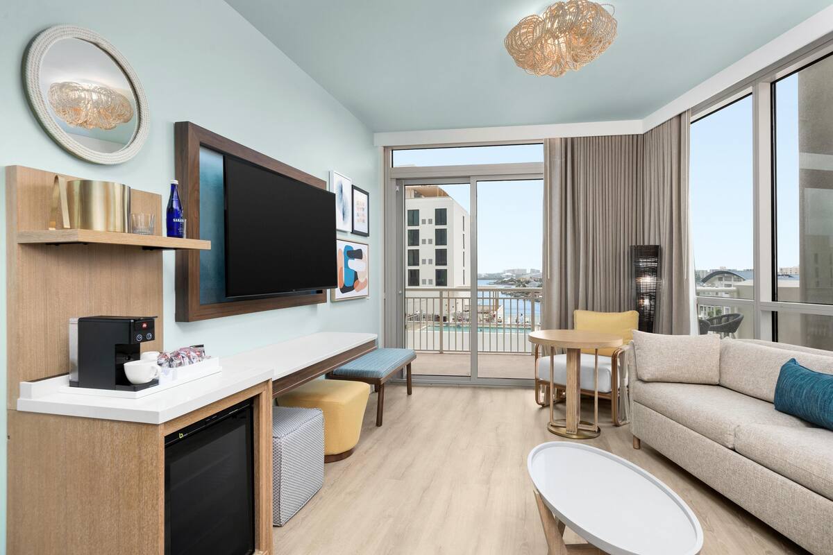 Een moderne hotelkamer met een flatscreen televisie, een koffieapparaat, een bank, een eettafel en een balkon met uitzicht op de stad.