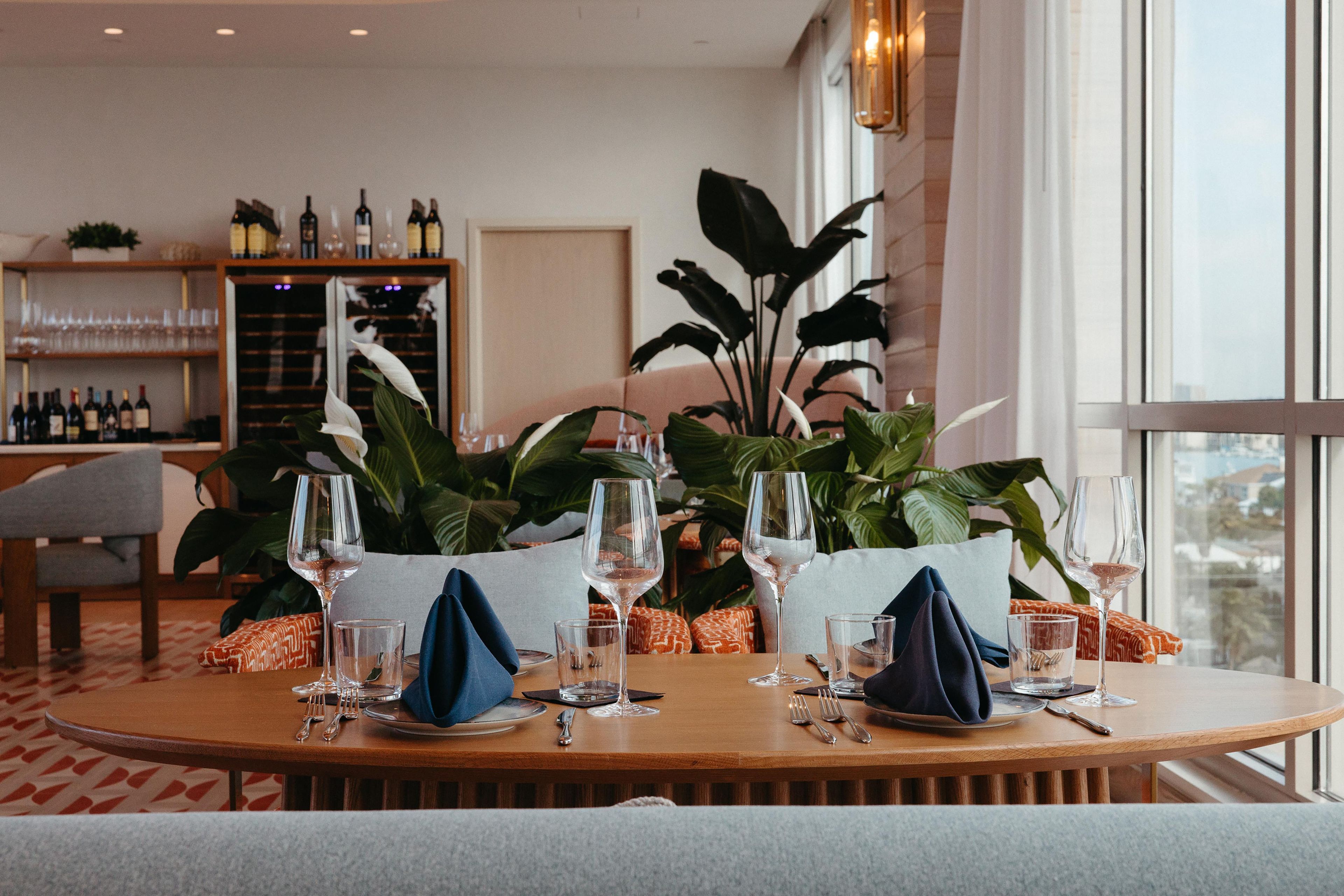 A dining area with a round table set for two, surrounded by large potted plants, with a wine rack in the background.