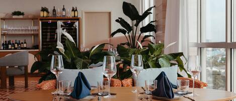 A dining area with a round table set for two, surrounded by large potted plants, with a wine rack in the background.