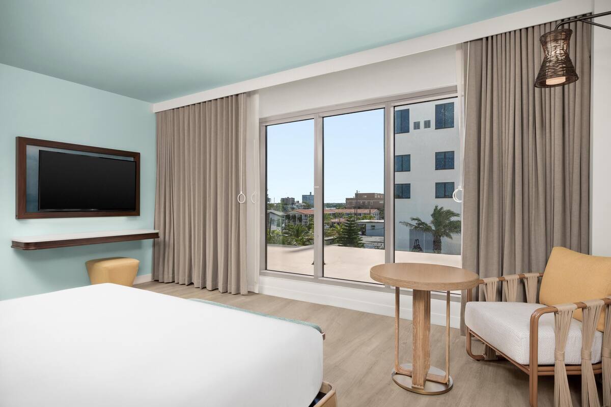Hotelkamer met een bed, televisie, klein tafeltje, stoel en uitzicht op de stad door grote ramen.