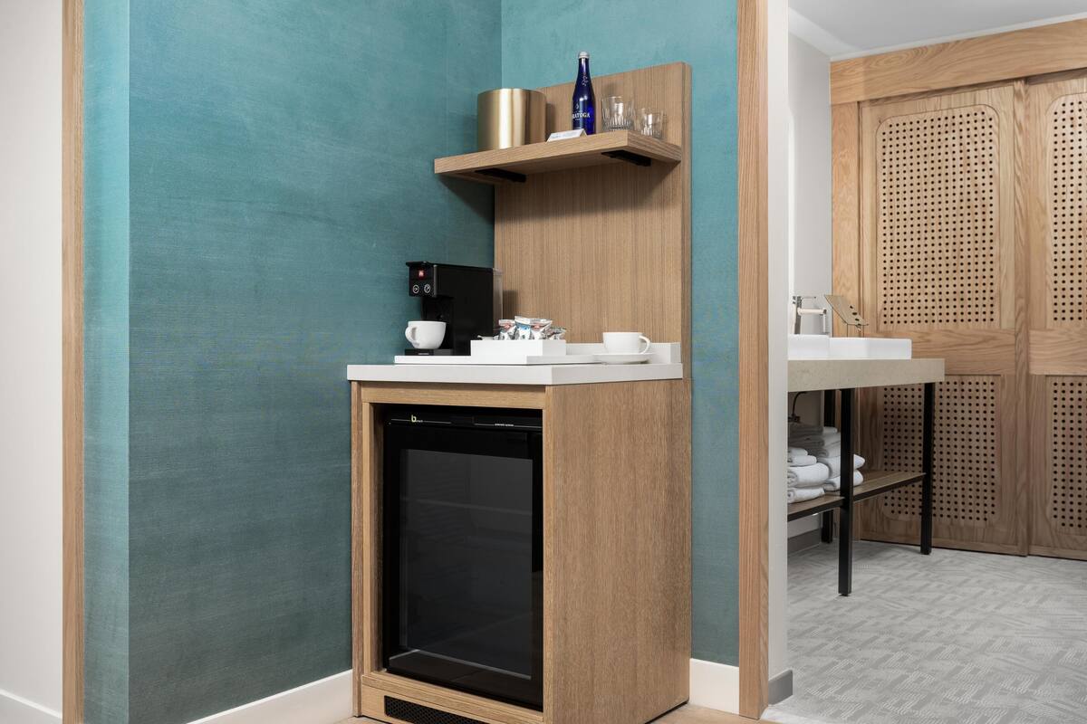 Een compact keukenhoekje met een koffiezetapparaat, een kleine koelkast en een houten plank met een fles en glazen.