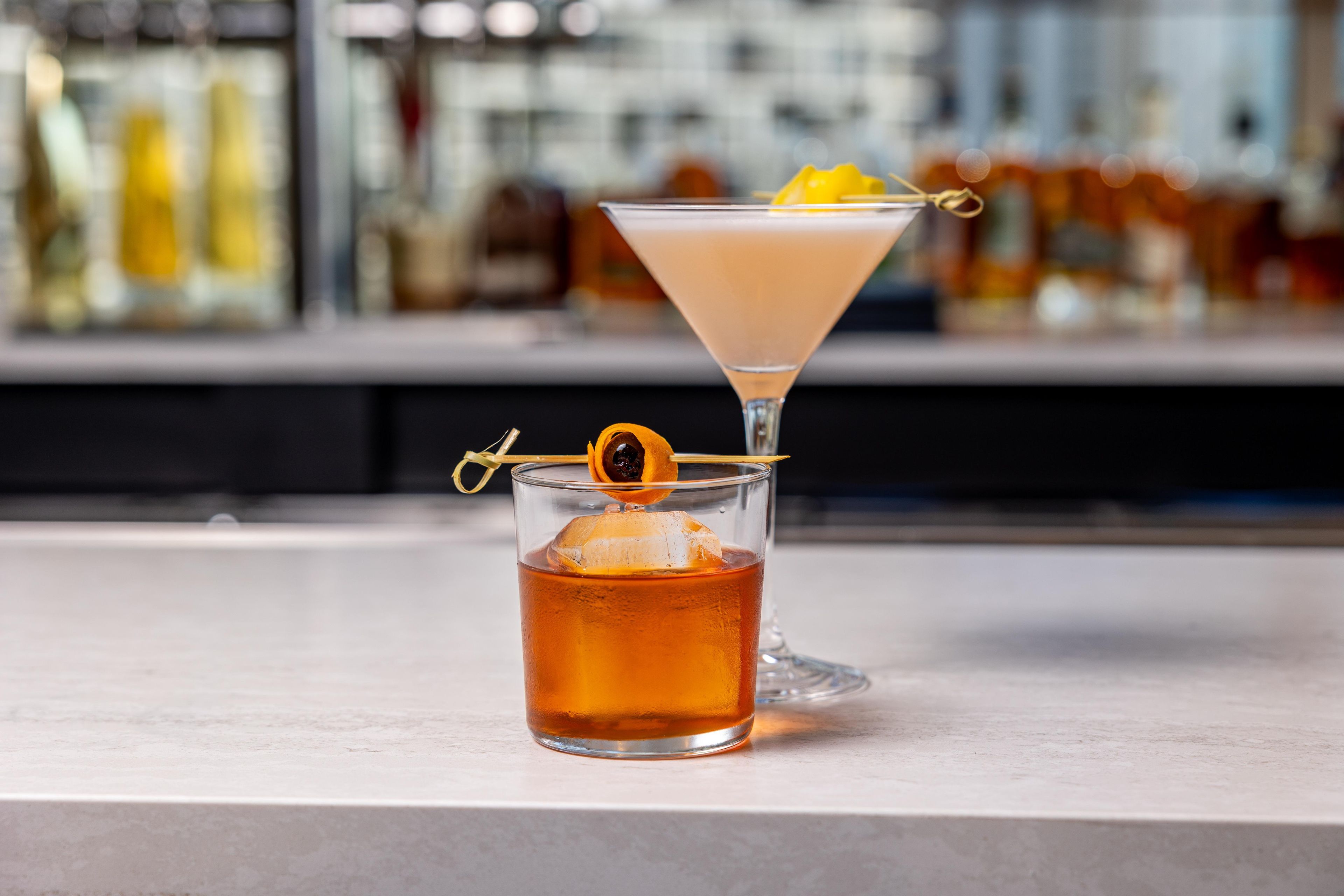 Two glasses of amber-colored beverages, one with a clear liquid and the other with a frothy top, on a bar counter.