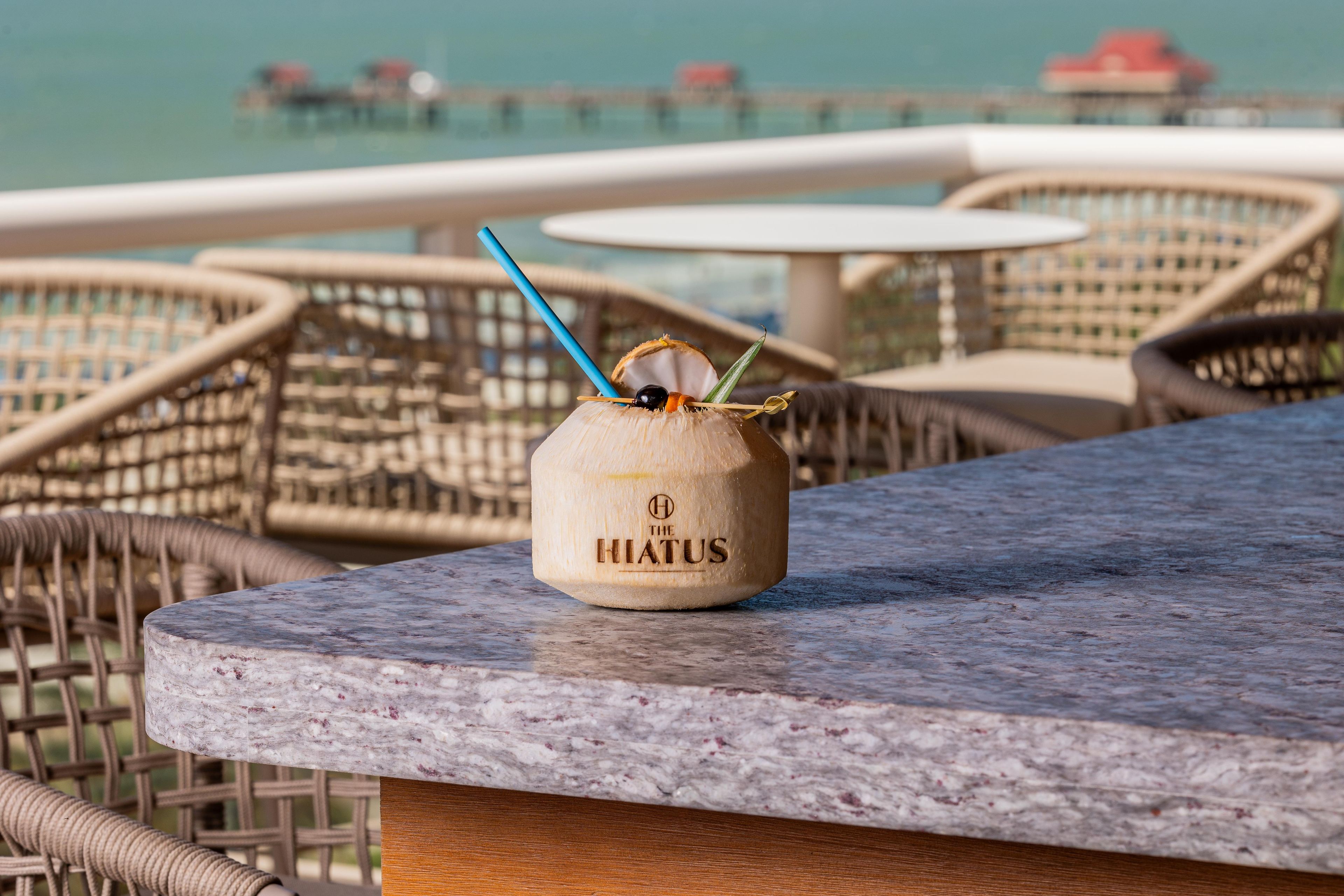 A coconut drink on a marble table with wicker chairs and a view of the sea in the background.