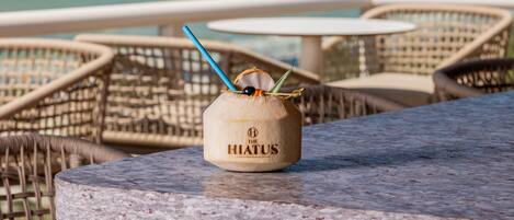 A coconut drink on a marble table with wicker chairs and a view of the sea in the background.