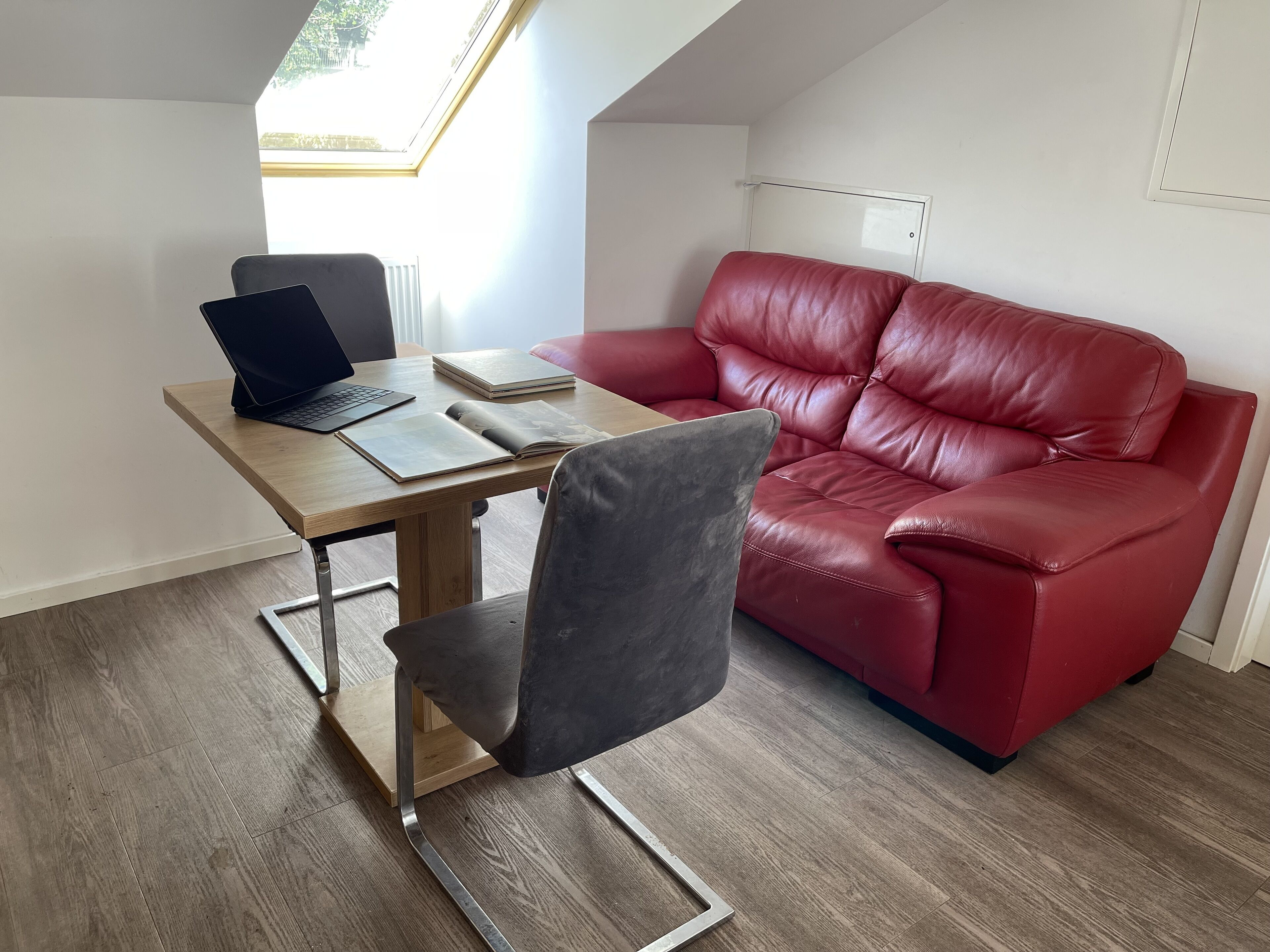 Una habitación moderna con un escritorio de madera, una computadora portátil, un libro abierto, un sofá de cuero rojo y un tragaluz.