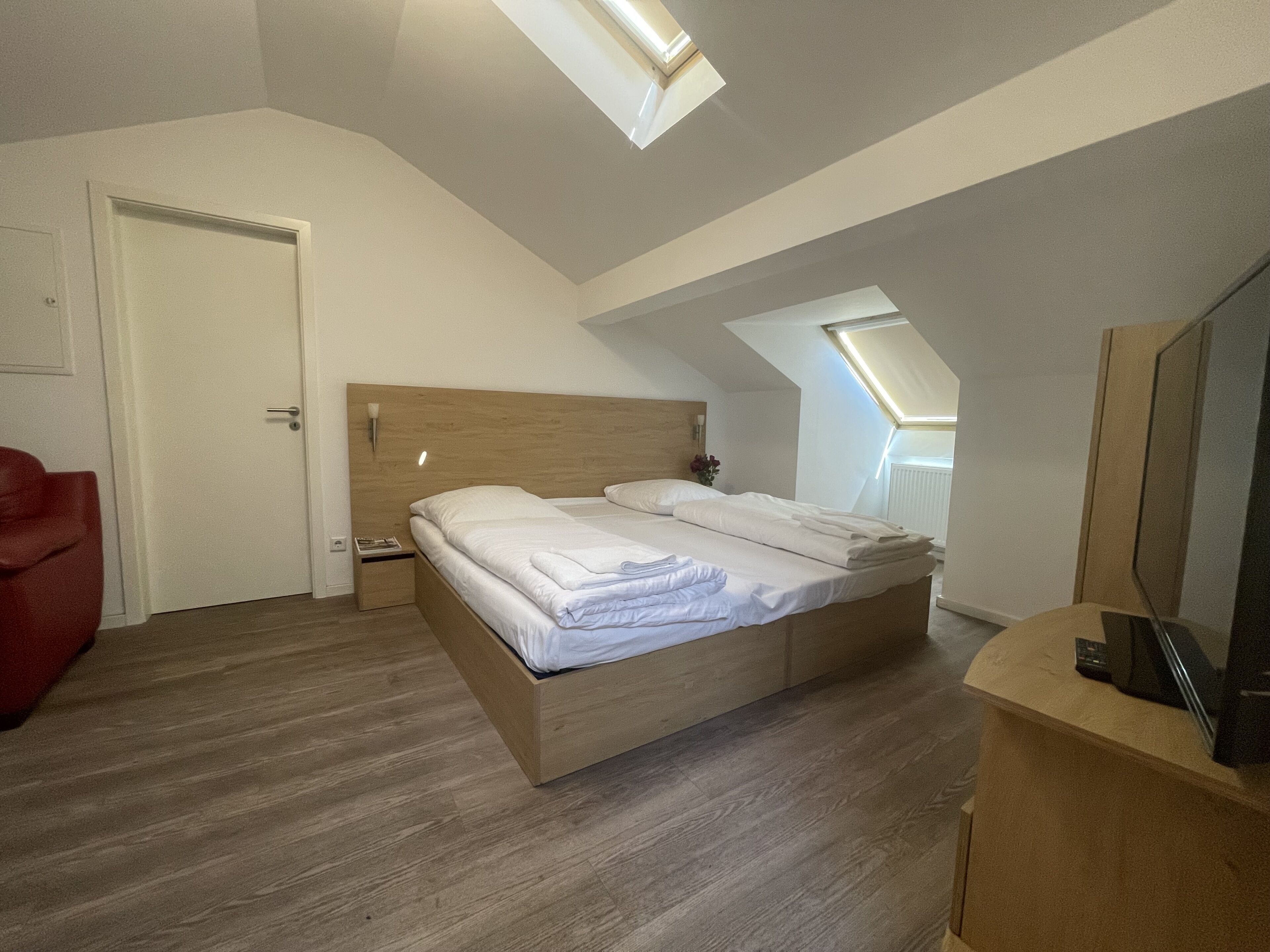 A bedroom with a wooden bed, a red chair, a nightstand, and a TV on a wooden stand.