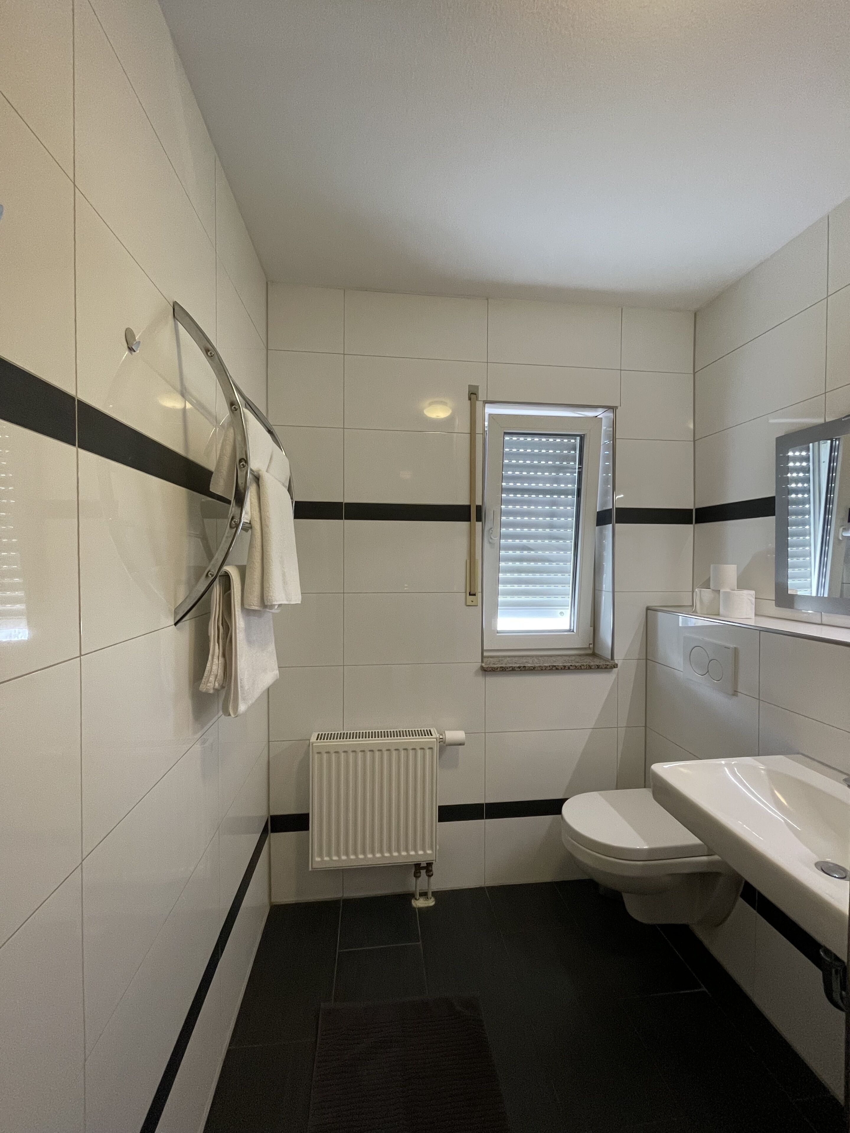 A bathroom with white tiled walls, a black stripe, a toilet, a sink, a towel rack, a window with blinds, and a radiator.