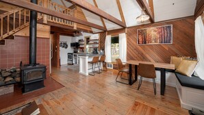 A spacious living area with a high ceiling, wooden beams, a fireplace, a dining area with a bench, and a kitchen in the background.