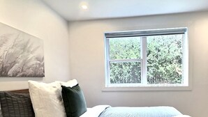 A bedroom with a bed, pillows, a blanket, a window with a view of trees, and a painting on the wall.