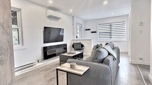 A modern living room with a grey sofa, a small coffee table, a flat-screen TV, and a window with blinds.